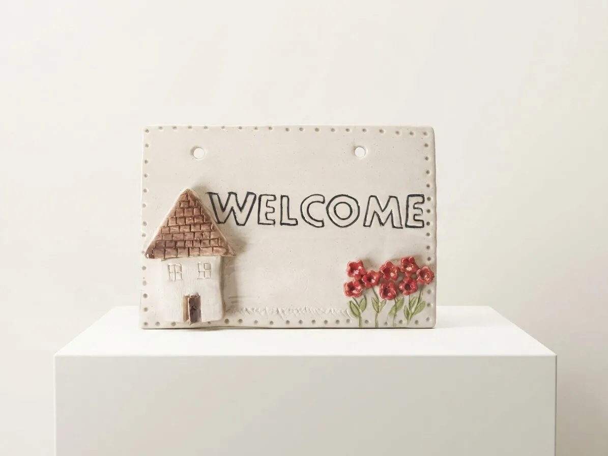 Welcome Tile — a cream ceramic wall plaque with a relief cottage, the word WELCOME and red poppies, edged with dots.