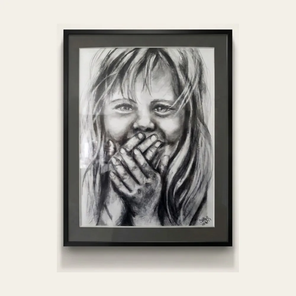Hidden Laughter — a charcoal drawing of a laughing girl covering her mouth with both hands, in a black frame.